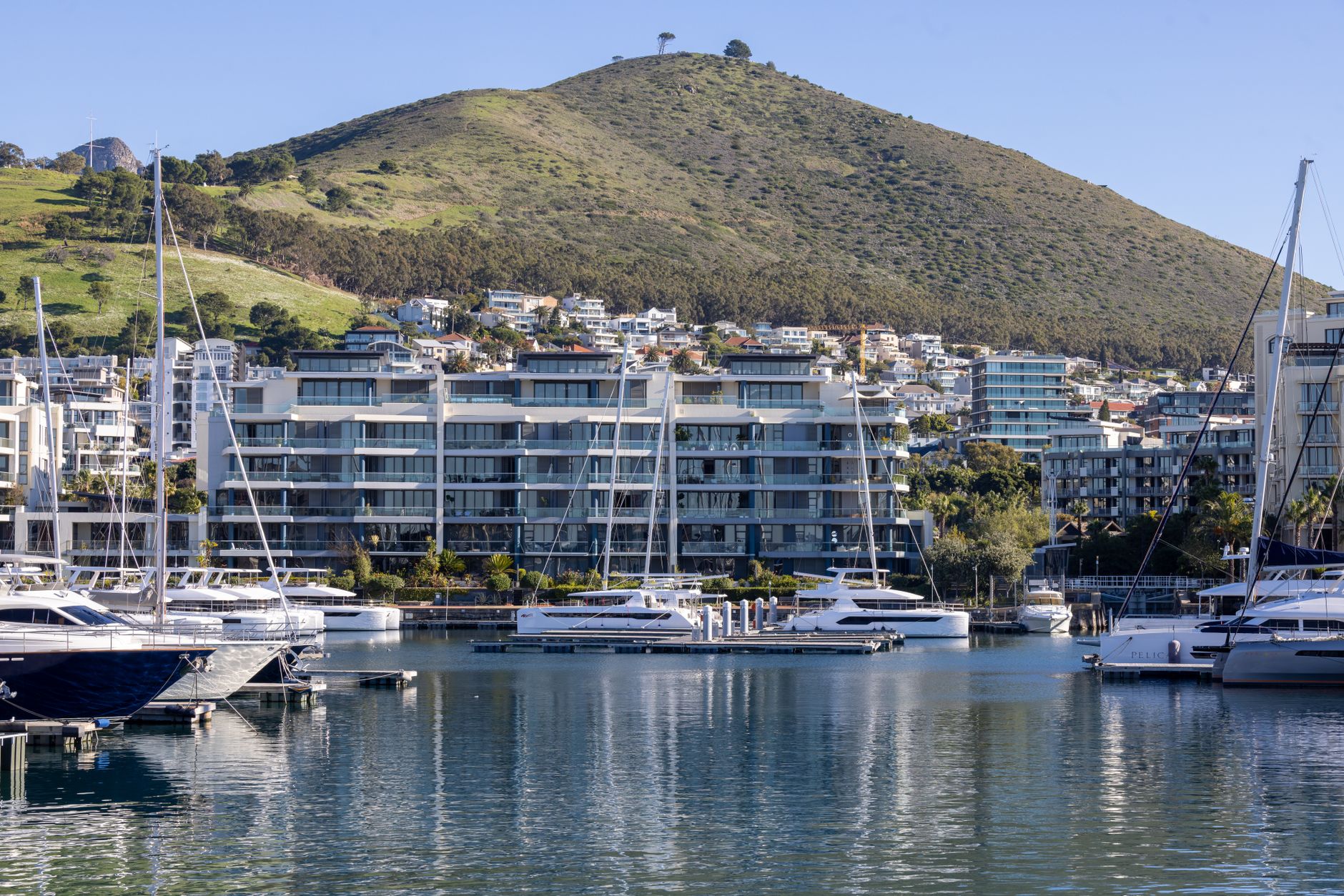 Waterfront view of luxury yachts in a scenic marina with hilly backdrop and modern apartments.